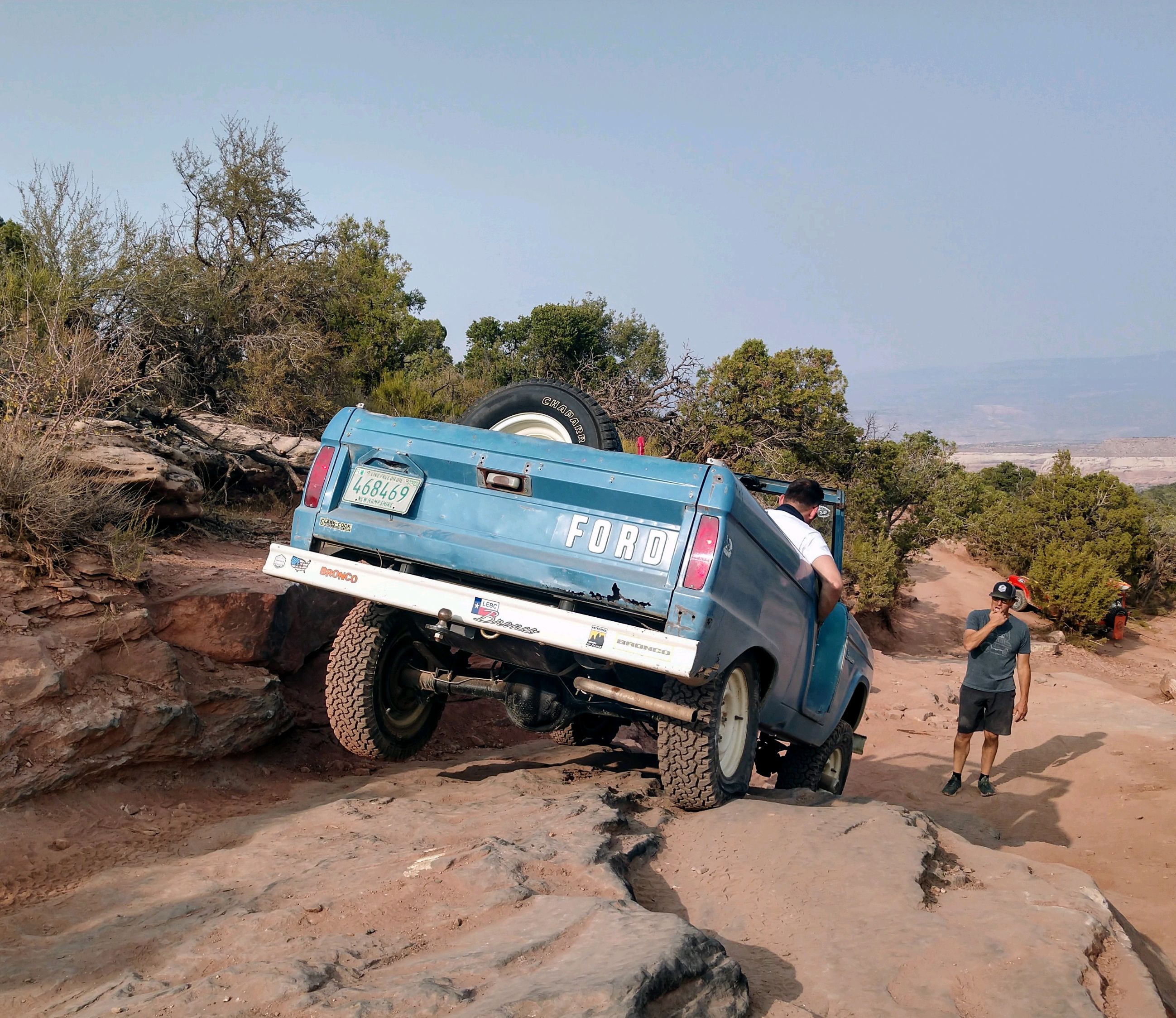 A blue vintage Bronco pictured from the back while driving over a rocky edge